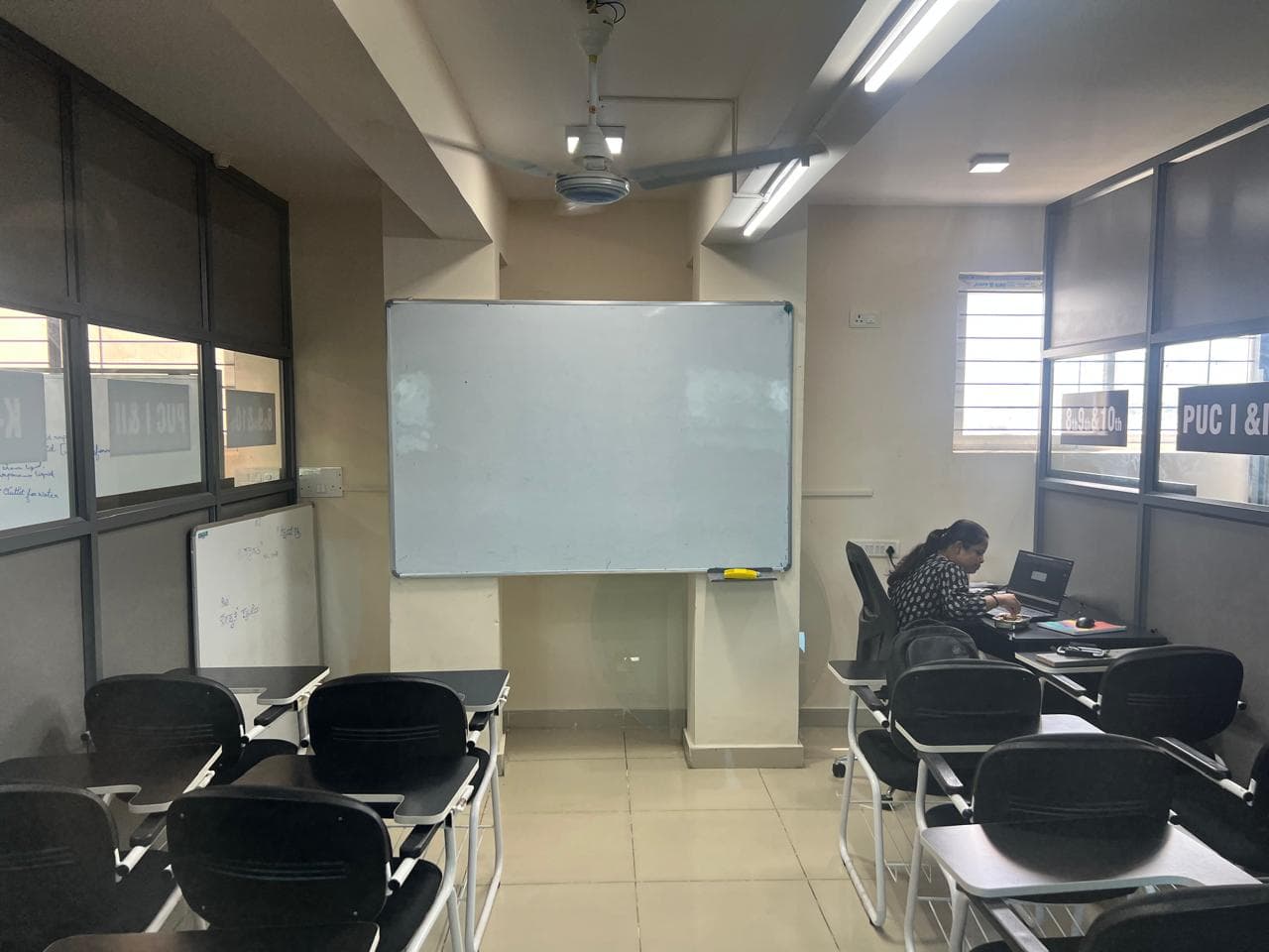 A teacher working at a laptop beside a whiteboard, projector and ceiling fan in a B-Cube classroom