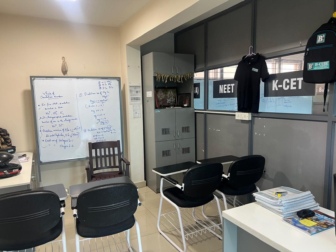 A B-Cube classroom with a whiteboard covered in worked equations, metal lockers, and NEET and K-CET signs on the glass partition
